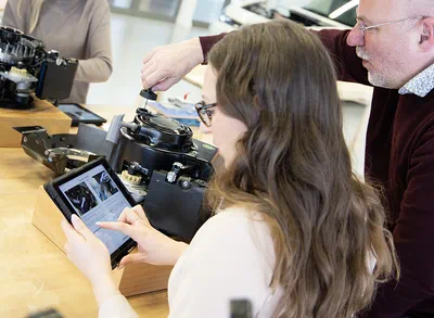 Eine junge Frau schaut auf einem Laptop während im Hintergrund ein Mann an einem elektrischen Gerät schaut