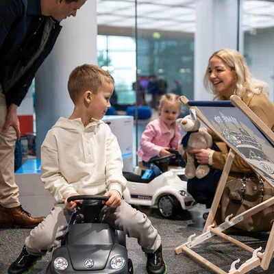 Zwei Kinder auf Volkswagen Bobby Cars und eine blonde Mutter, die einen Teddy in der Hand hält