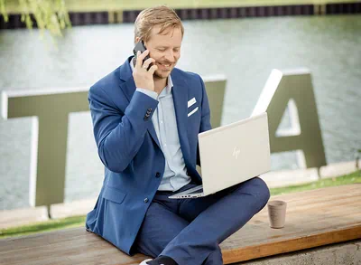 Ein Mann sitzt auf einer Bank am Wasser, hält einen Laptop und spricht am Telefon